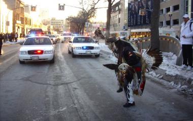 Photo: Native American dressed in traditional garb in front of a flashing cop cars