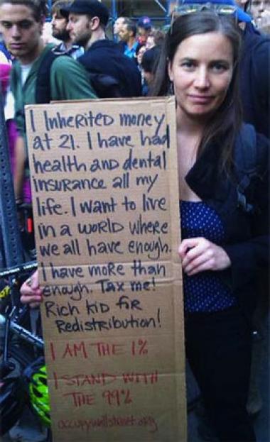 Woman holding sign: I inherited money at 21. I have had health and dental insurance all my life. I want to live in a world where we all have enough. I have more than enough. Tax me! Rich kid for redistribution. I AM the 1%. I stand with the 99%