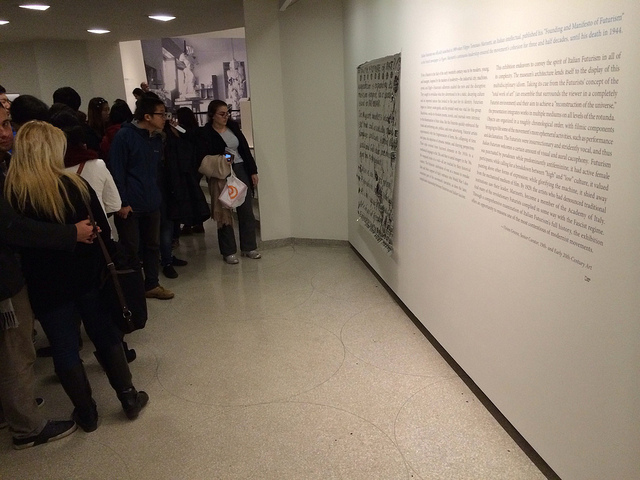 Museum visitors reading the manifesto tacked to the wall beside the introductory text to the Futurism exhibition.